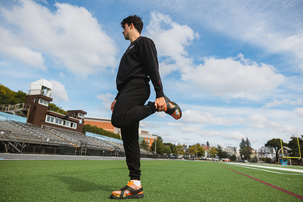 Athlete stretching on turf while wearing CUVS CV1 Blackout spike covers over orange track spikes.